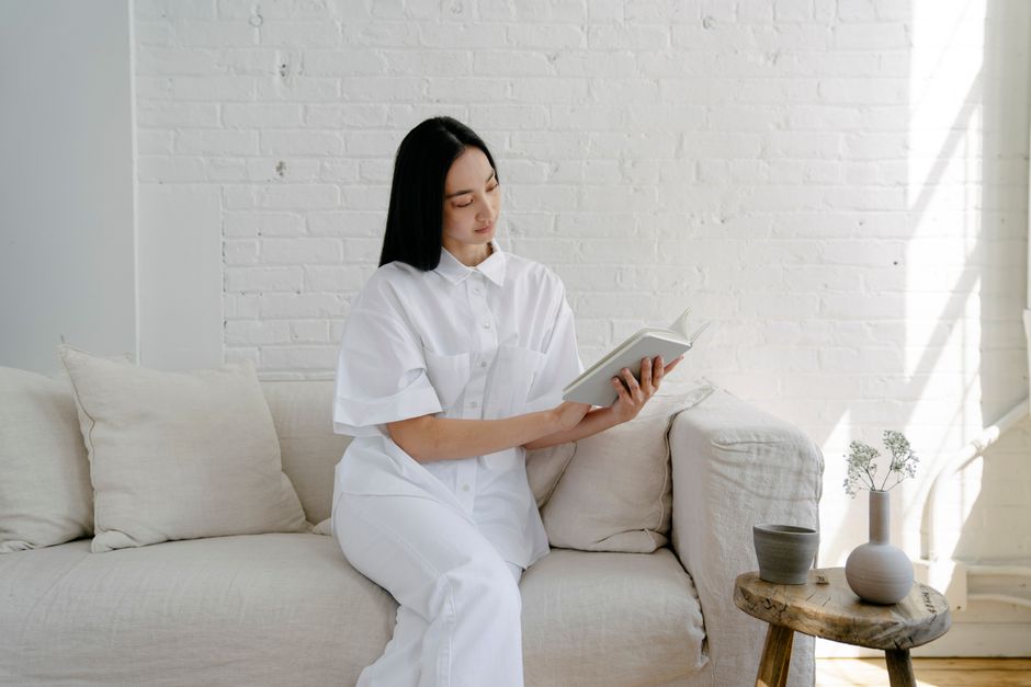 Young ethnic female in white clothes sitting on sofa and reading book in cozy room