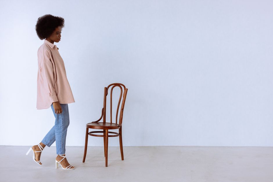 A woman in casual attire walks by a wooden chair, against a plain background.
