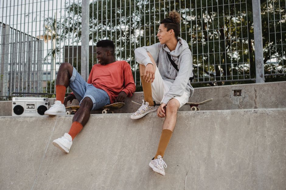 Full body of positive diverse male skaters resting on ramp in skate park with boombox and listening to music