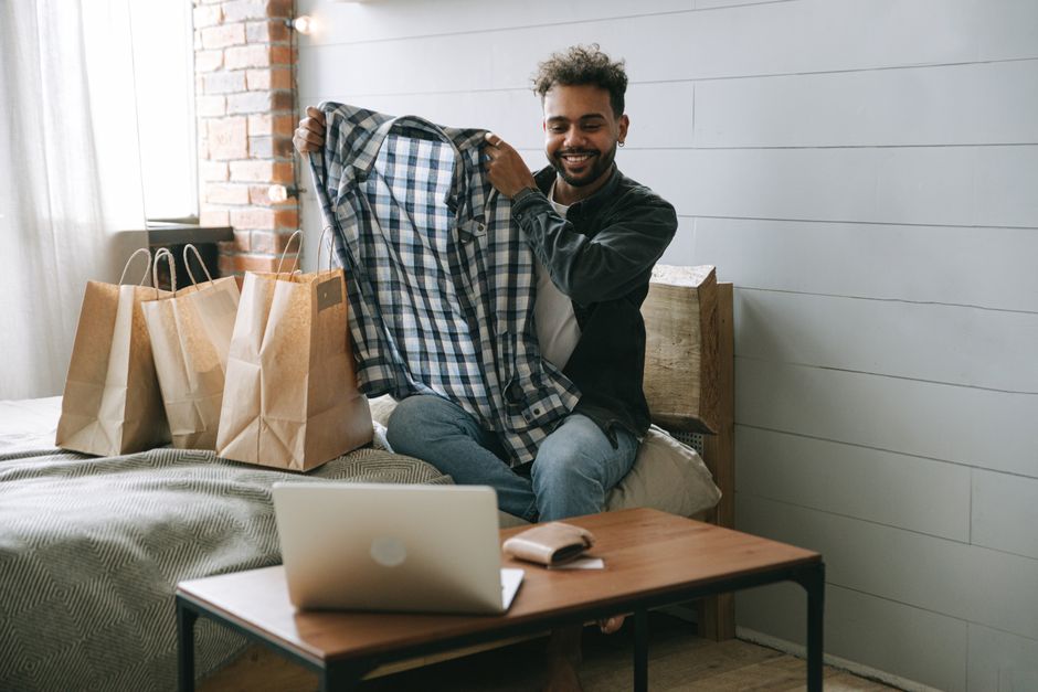 Smiling man excitedly unpacking new clothes from shopping bags in his cozy bedroom setting.