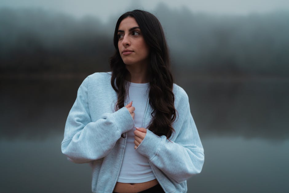 A young woman wearing a casual jacket stands against a serene, foggy lake background.