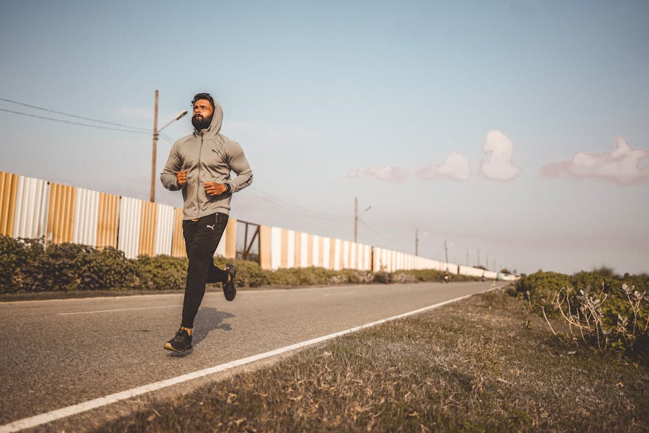 A bearded man in a hoodie jogs along an empty road under a clear sky.
