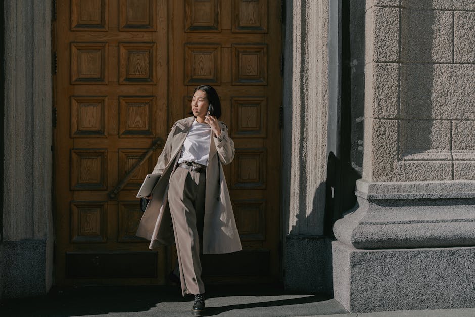 Trench-coated woman standing confidently in front of a wooden door during the day, using a smartphone.
