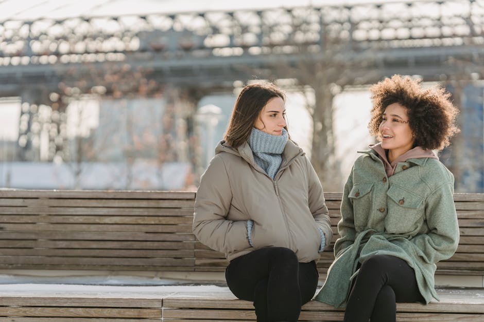 Cheerful multiracial female friends in warm clothes sitting on wooden bench while spending time together on street in winter day