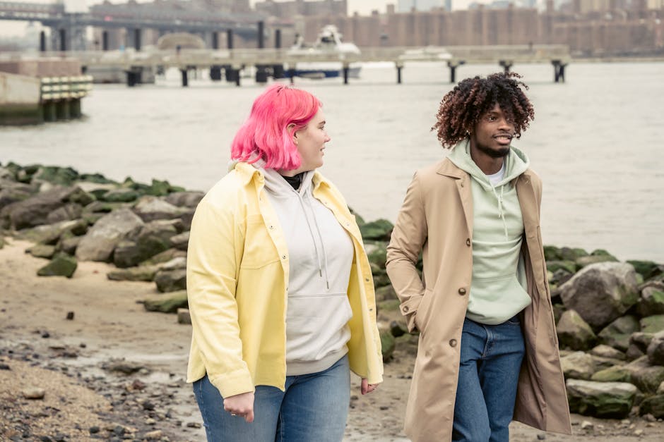 Content multiracial couple in outerwear strolling on embankment with stones near calm river against bridge and residential buildings in city