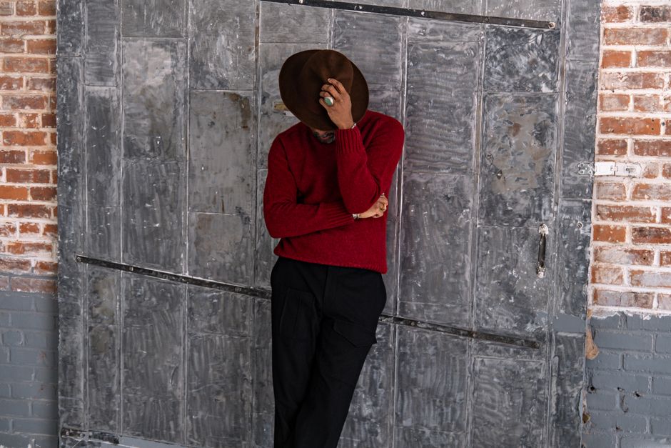 Stylish man leaning against a textured wall, wearing a brown hat and red sweater.