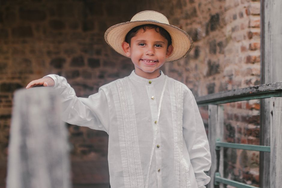 Smiling boy in traditional white clothing and hat standing outdoors.