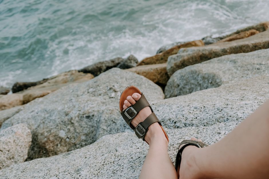 A person relaxes on a rocky seashore wearing sandals, enjoying the coastal view.