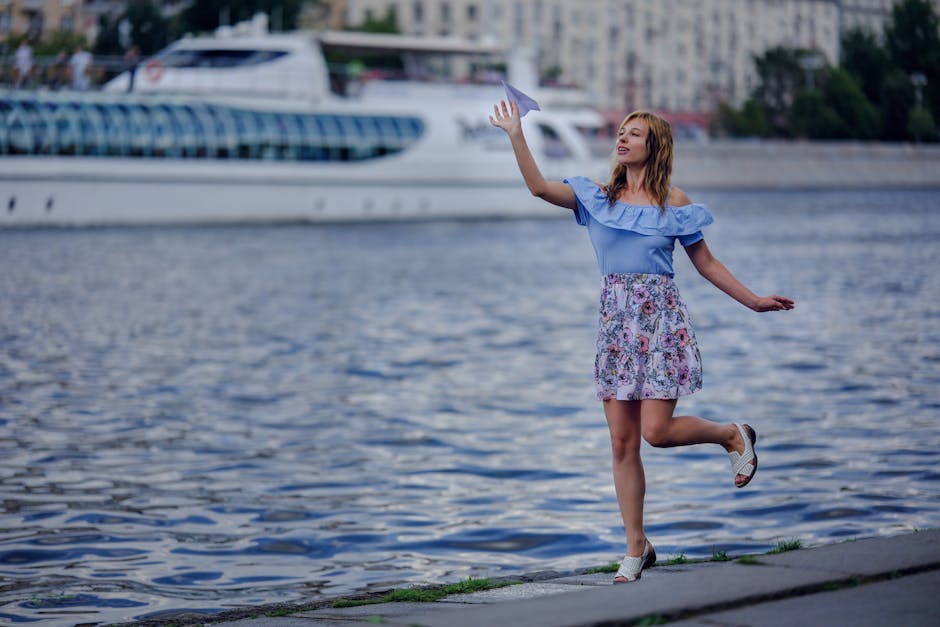 A woman stands by a river with a paper plane, posing in an urban environment with a ship in the background.
