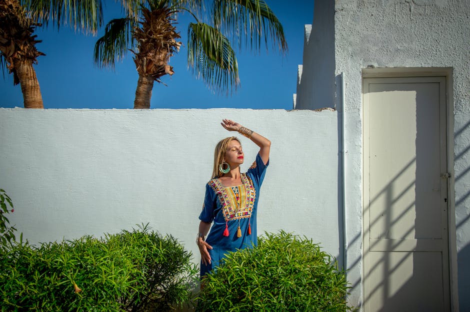 Fashionable woman in blue dress with arm raised, posing outdoors against a white wall and palm trees.