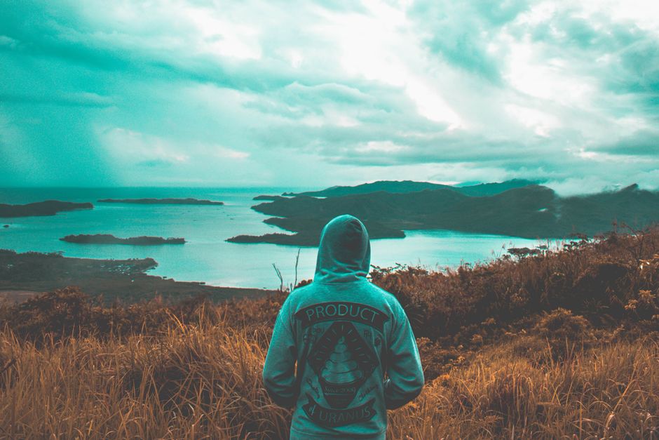 Person in a hoodie overlooking a beautiful coastal vista with islands under a cloudy sky.