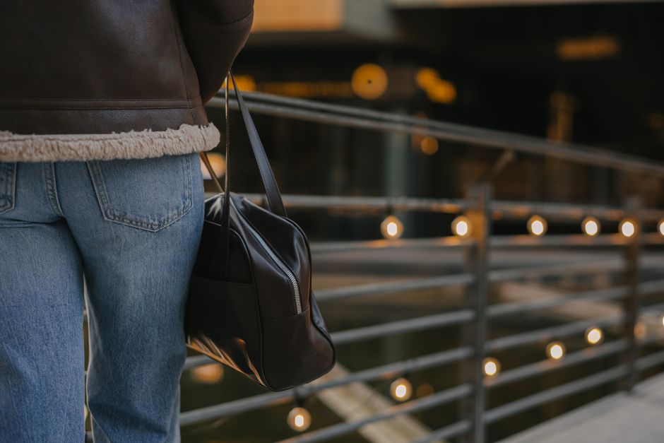 Casual scene of a person in jeans and a leather jacket holding a fashionable leather bag on a lit bridge.