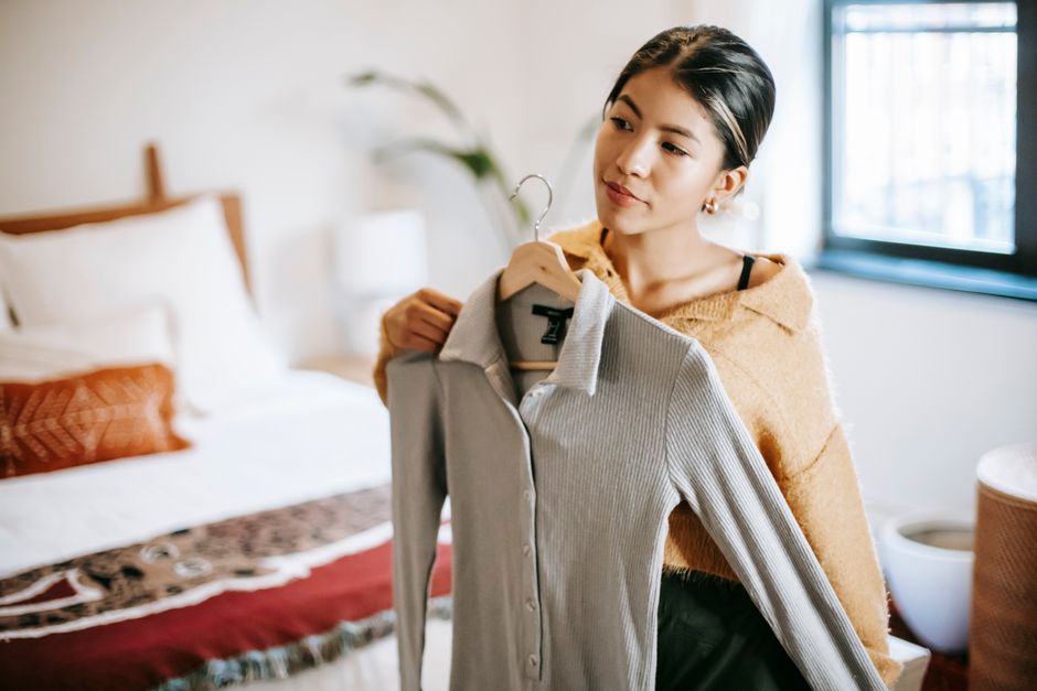 Ethnic female trying on jacket with buttons on hanger while choosing outfit at home