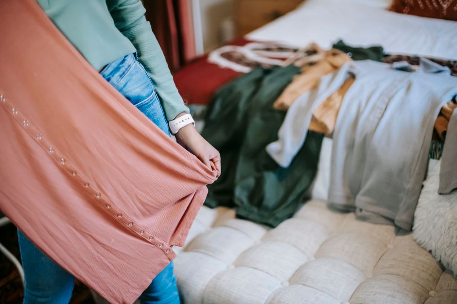 Crop anonymous woman in casual clothes demonstrating pink dress while selecting clothes while standing in bright bedroom near couch and bed with wardrobe at home