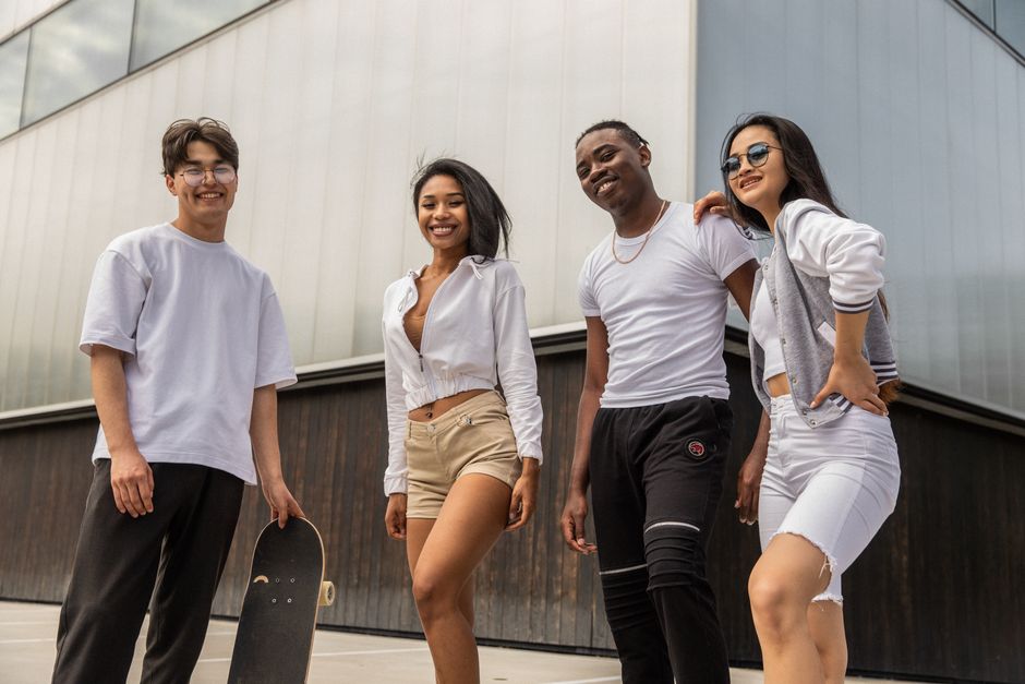 Low angle of cheerful young multiracial male and female friends in activewear standing near modern building and looking at camera after riding skateboards on street