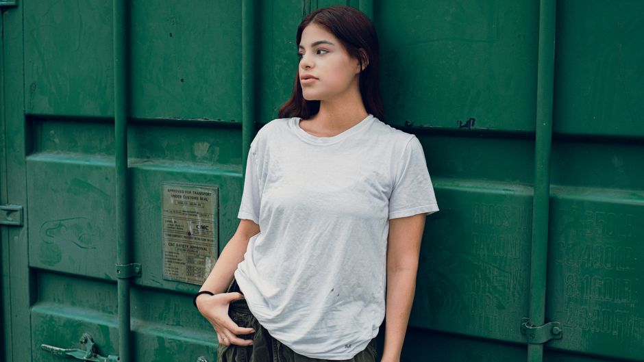 Casual portrait of a woman in a white t-shirt leaning against a green container outdoors.