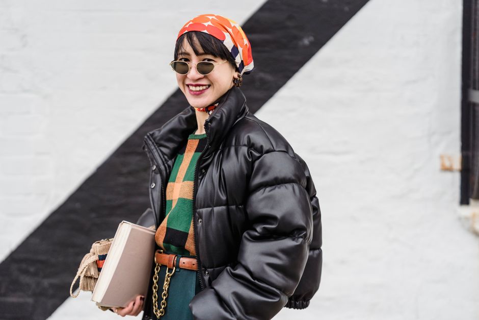 Cheerful Asian female in trendy headscarf and sunglasses looking at camera while standing near wall of building with notebook in hand