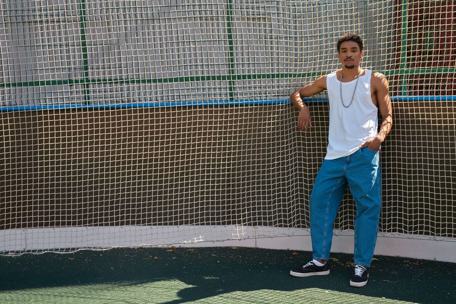 A young man poses in casual streetwear, leaning against a net outdoors in daylight.