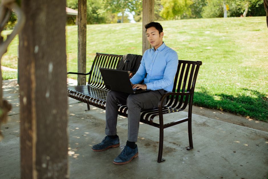 Asian businessman in corporate attire working on laptop outdoors in a sunny park setting.
