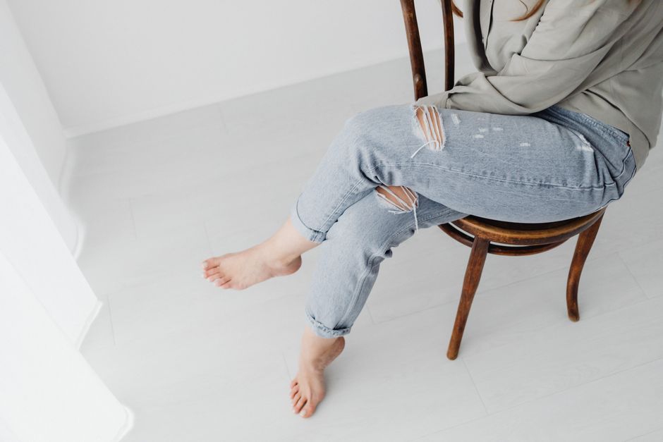 Woman sitting on wooden chair with crossed legs wearing ripped jeans in minimalistic room.