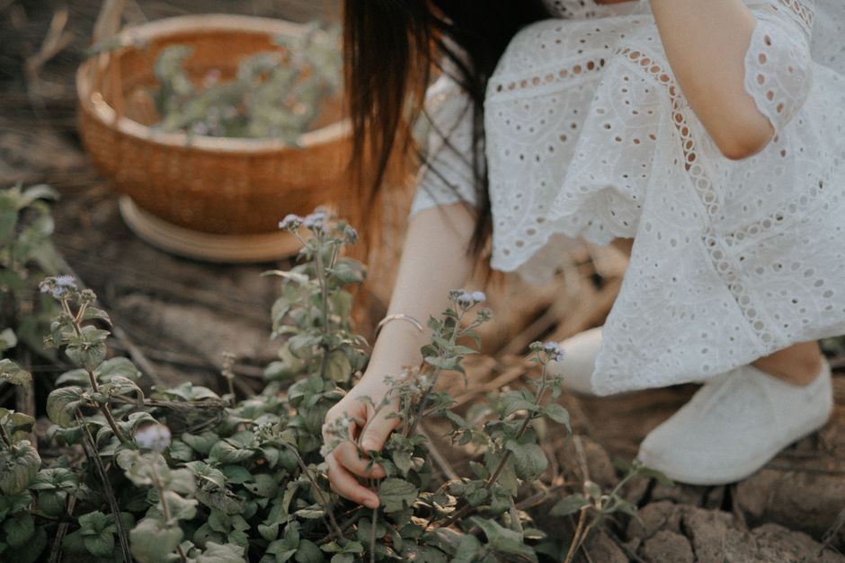 A woman in a white dress picking wildflowers in a natural outdoor setting.
