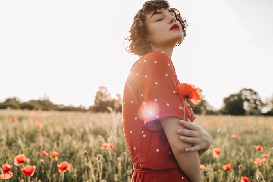 A serene portrait of a woman in a red dress holding a poppy in a sunlit field.