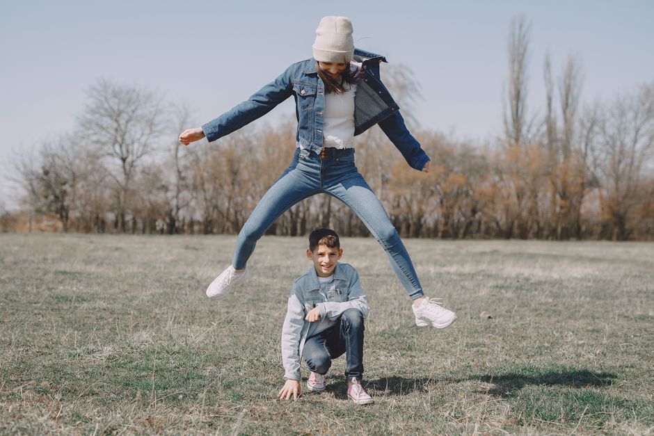 Delighted young sister and brother in trendy clothes jumping across each other while resting together on lawn in park at sunny spring day