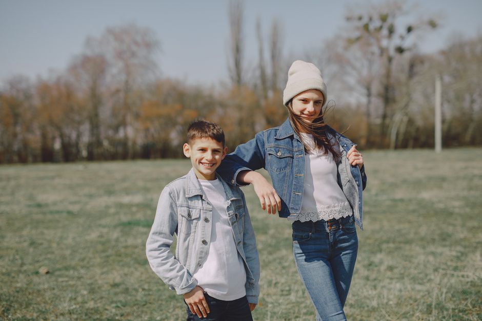 Cheerful brother and sister in jean outfit smiling and cuddling while looking at camera in countryside at daytime