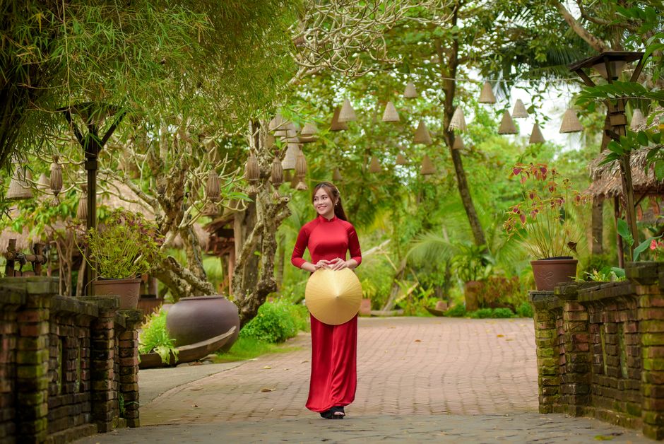 Woman in traditional red ao dai holding conical hat in lush garden setting, exuding cultural charm.