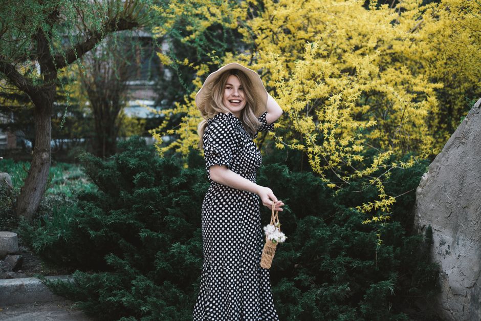 A woman in a polka dot dress poses happily with a straw hat in a vibrant spring garden.