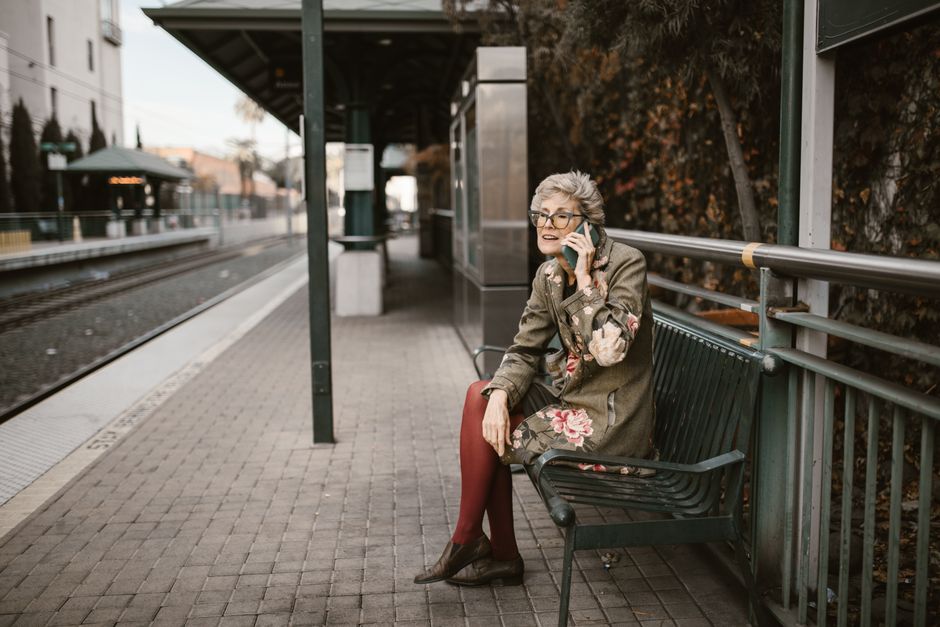 Elderly woman sitting on bench at outdoor train station using smartphone.