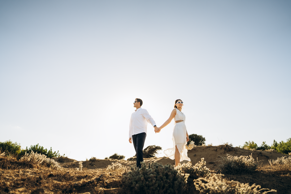 A stylish couple walks hand in hand on a sunny day in Antalya, Turkey, amidst a natural landscape.