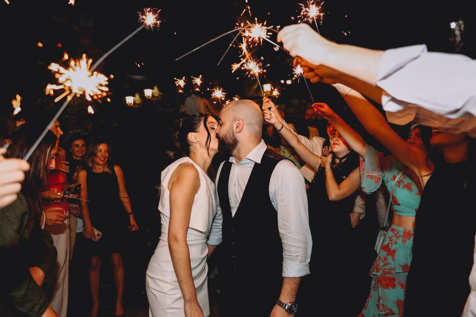 Couple celebrates wedding with sparklers surrounded by friends at night.