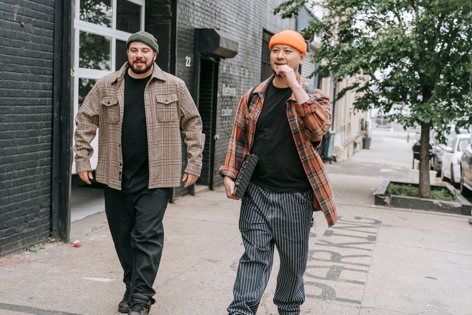 Two men in beanies and plaid shirts walking on a city sidewalk, showcasing urban fashion.