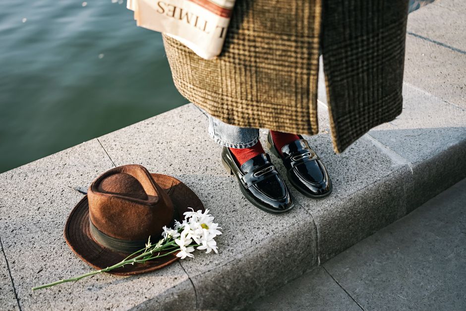 Chic fashion ensemble featuring patent loafers, hat, and newspaper by the water.