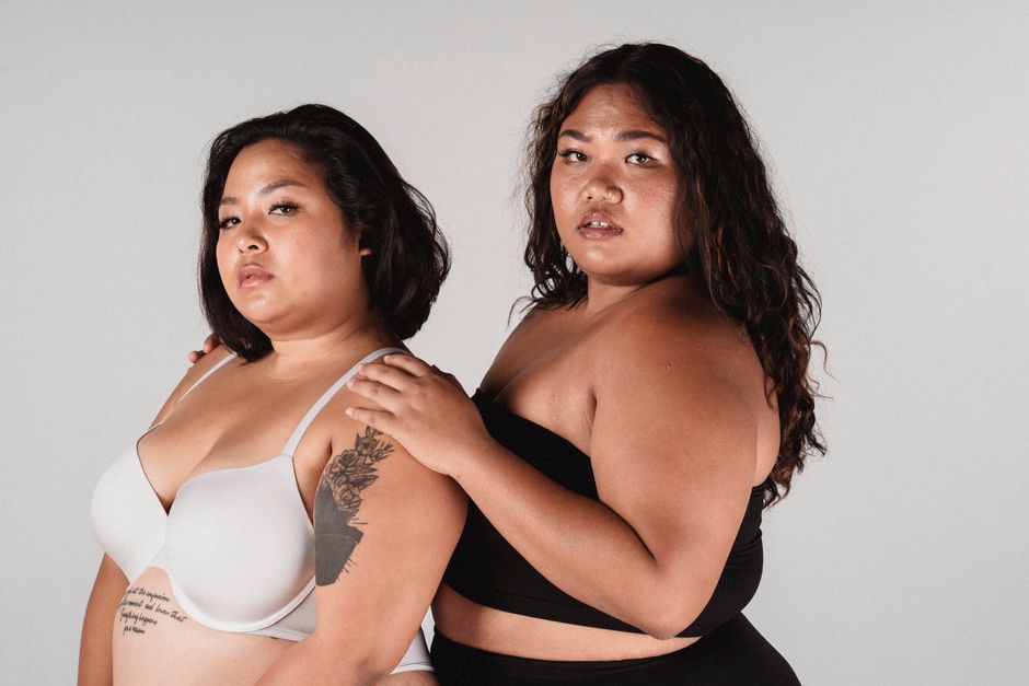 Side view of plus size ethnic woman leaning on shoulder of tattooed female in underwear on white background